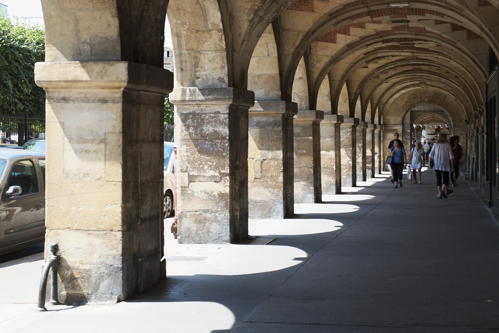 The vaulted stone arcade of the Place des Vosges with people walking in the shade